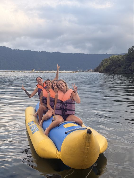 Four friends in life jackets cheering on a yellow banana boat on calm water surrounded by green hills