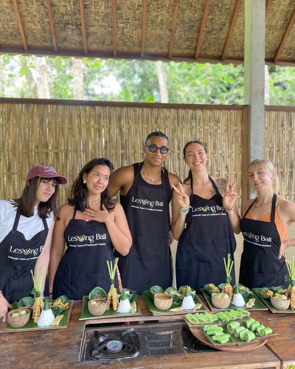 Five people in matching aprons standing behind dishes served on banana leaves at a cooking class