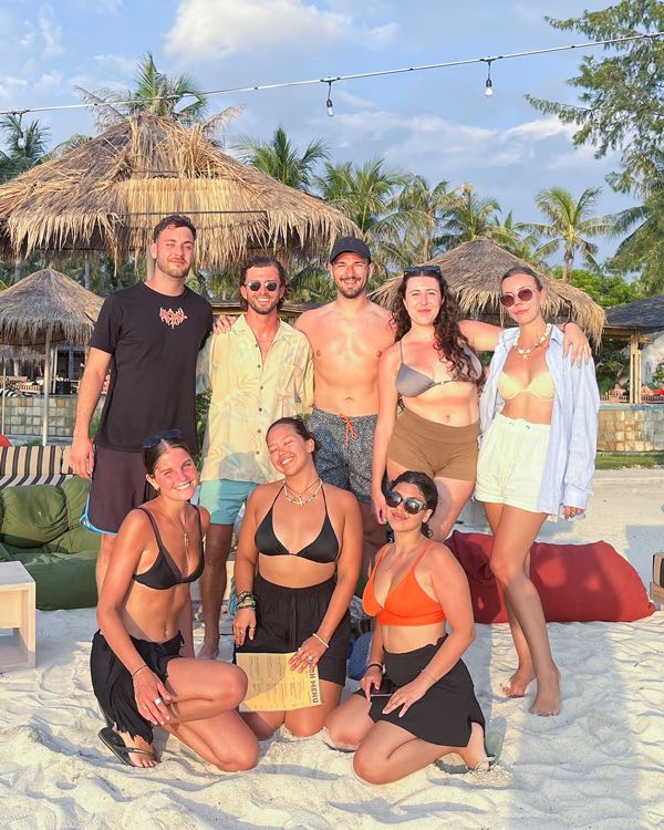 A group of friends in swimwear posing on white sand under a thatched beach bar