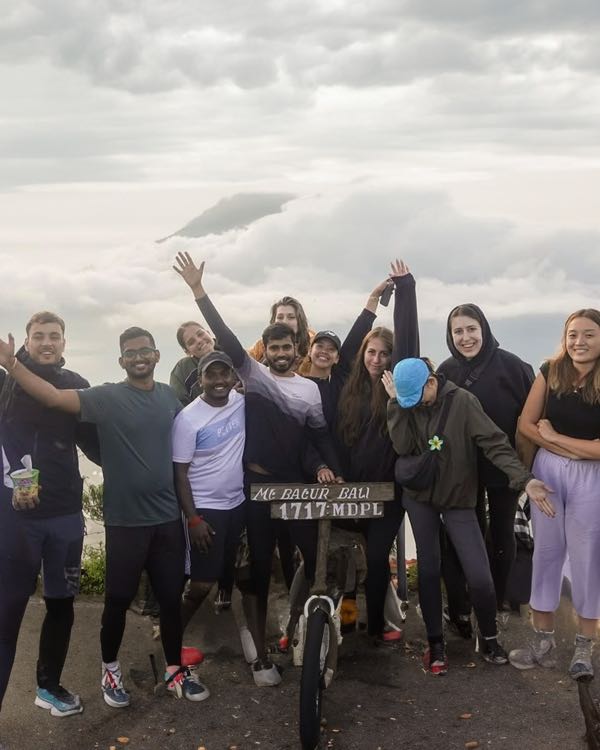 A group cheering with arms raised next to the Mount Batur summit sign, with clouds behind them