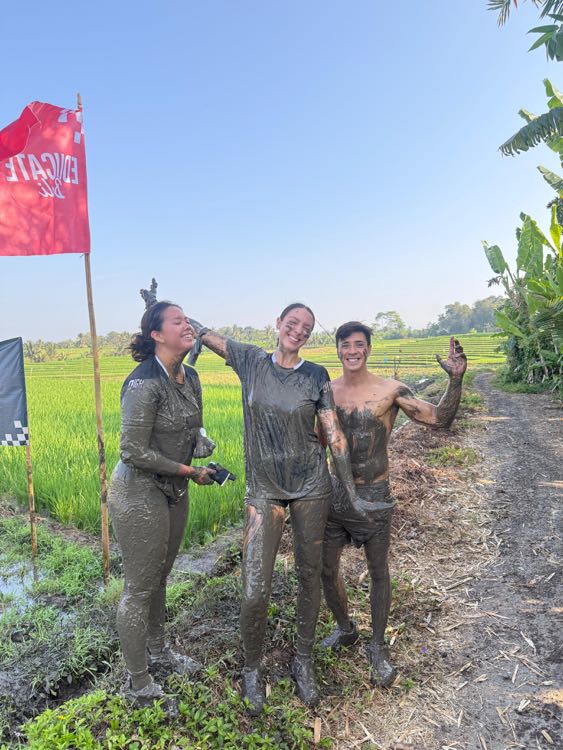 Three people covered head to toe in mud, standing in a rice field next to a red flag