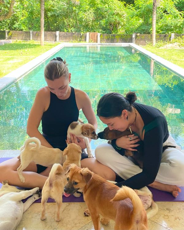 Two women on yoga mats beside a pool, surrounded by puppies
