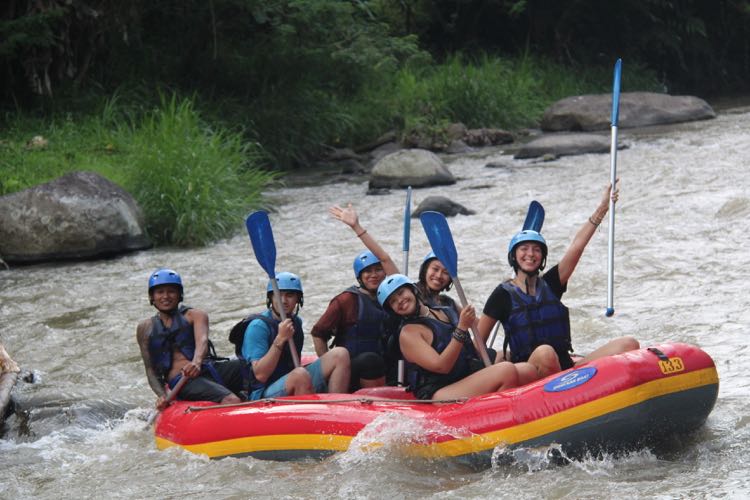 Six people in helmets and life jackets cheering with paddles raised in a red raft on a jungle river