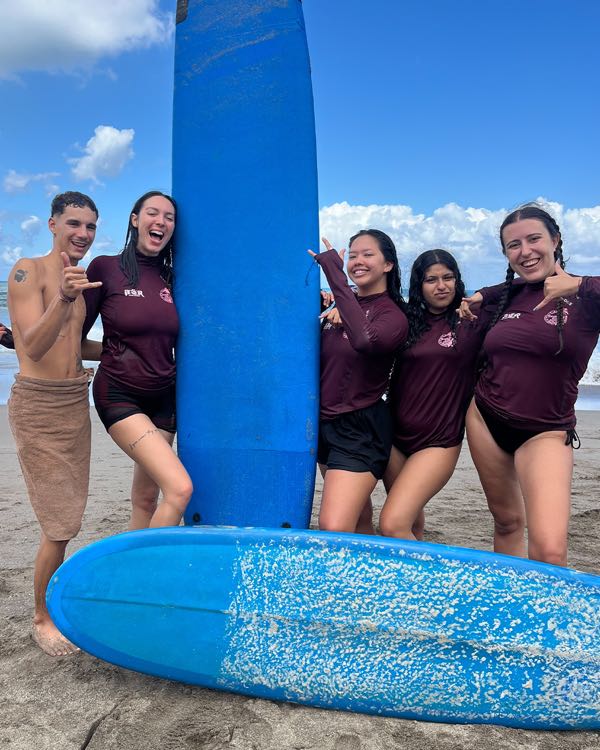 A group in maroon rash guards laughing on the beach next to large blue surfboards